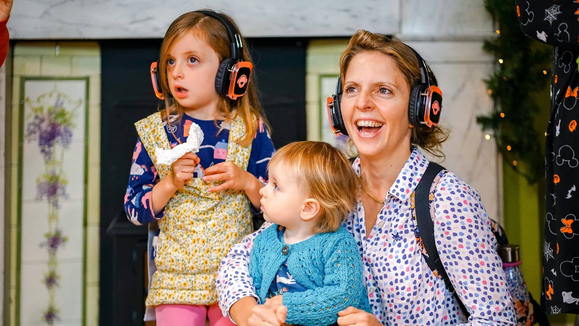 A woman with brown hair sits with her two children with a big smile on her mouth. The mother and her eldest daughter are wearing headphones.