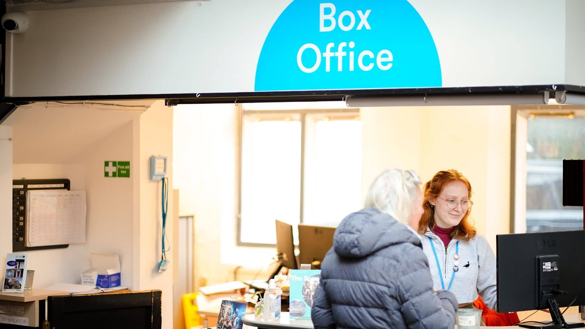 Two women, one with ginger hair with glasses and one with white hair are chatting at the box office at The Spring.