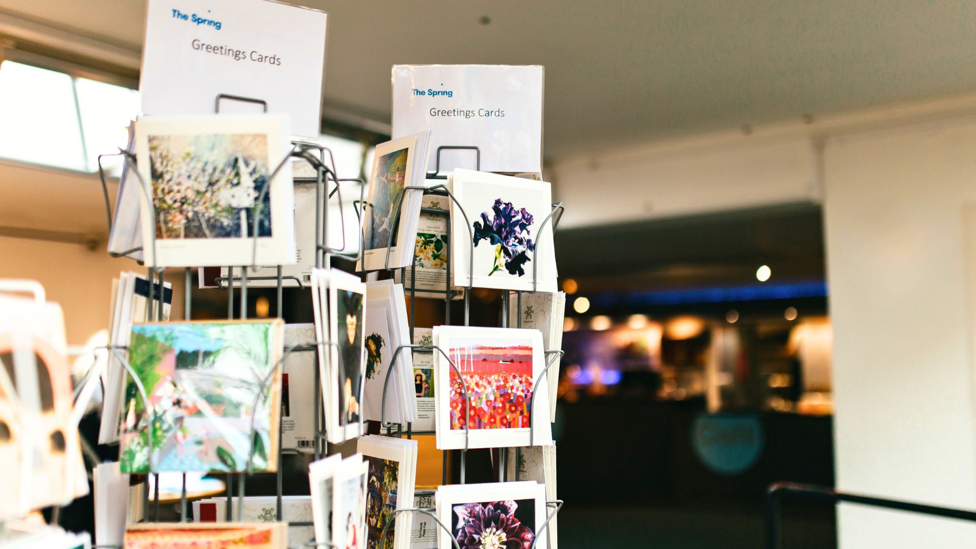 A display rack of greeting cards in a shop, labelled "The Spring Greetings Cards. The cards feature various colourful designs, including floral patterns and scenic artwork. The background is softly blurred, showing an interior space with warm lighting and indistinct details of a counter or seating area.