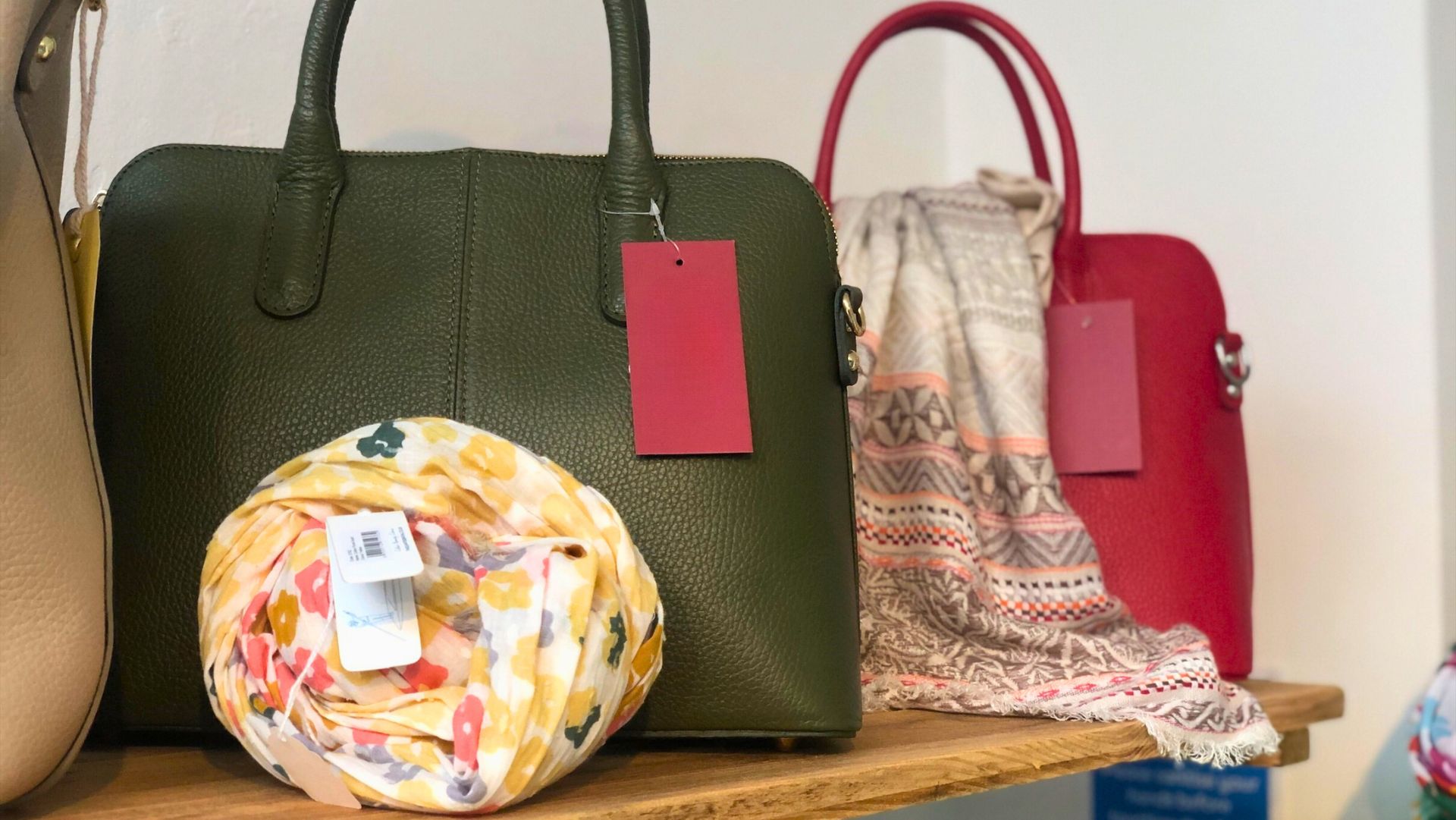 An arrangement of handbags and accessories on a wooden shelf. A green handbag with a red tag is paired with a rolled-up yellow and floral-patterned scarf, while a red handbag is displayed alongside a folded beige and geometric-patterned scarf. The background is simple and neutral, drawing attention to the products.