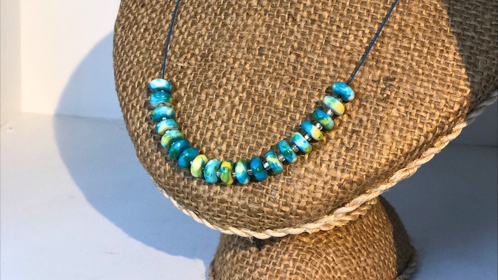 A close-up of a colourful necklace displayed on a textured burlap stand. The necklace features vibrant blue, green, and yellow beads strung on a thin wire, creating a bright and eye-catching design. The simple background highlights the handmade jewellery.