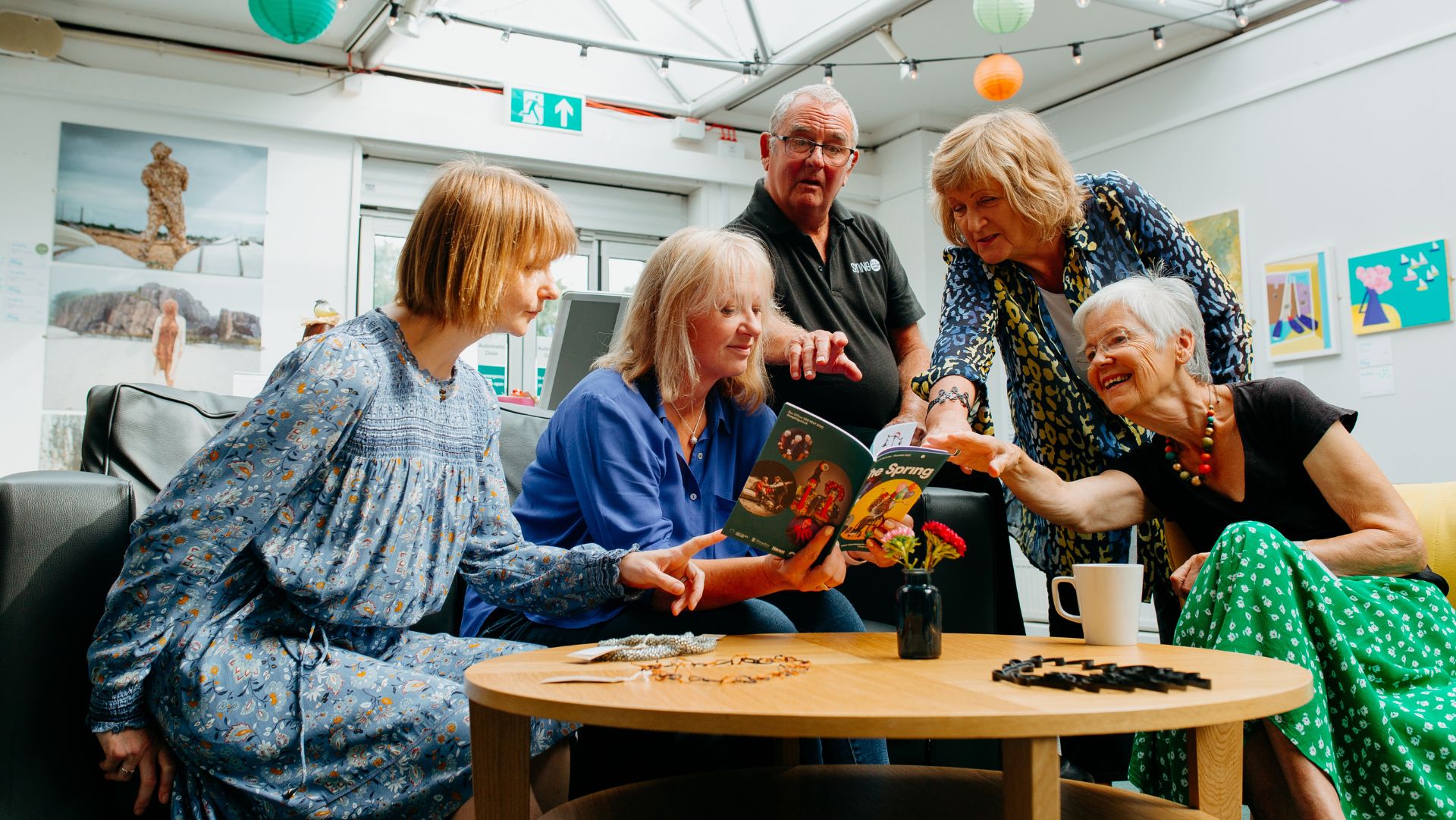 A group of five people are gathered in a bright, cosy community space, looking at a booklet titled "The Spring". Three women sit on a sofa and armchair, while a man and another woman stand behind them. A coffee table in the foreground holds a red flower in a vase, a mug, and craft materials. The atmosphere is warm and collaborative.