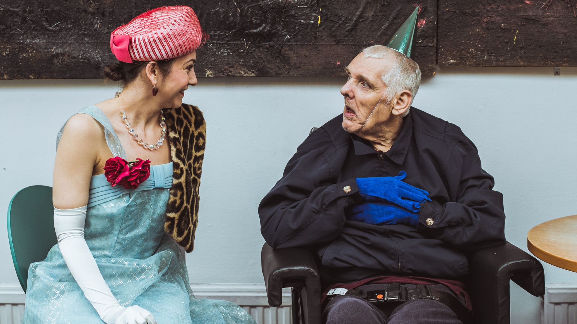 A woman with brown hair and wearing a traditional outfit with a pink bowed hat, light blue dress with a bouquet of red flowers on and long white gloves is looking at a man with gray hair sat in a wheelchair wearing a green party hat and blue gloves