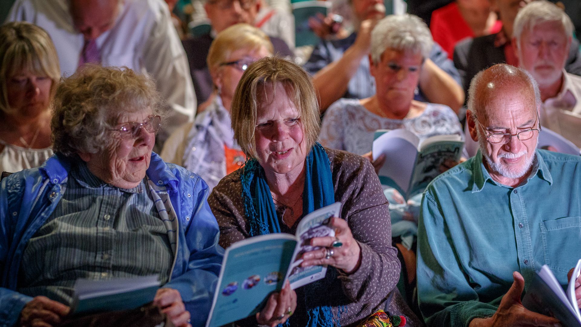 An audience of elderly people sitting and reading a programme. A woman in the front row with brown hair has the book open to show another woman with gray hair and glasses the content of the book