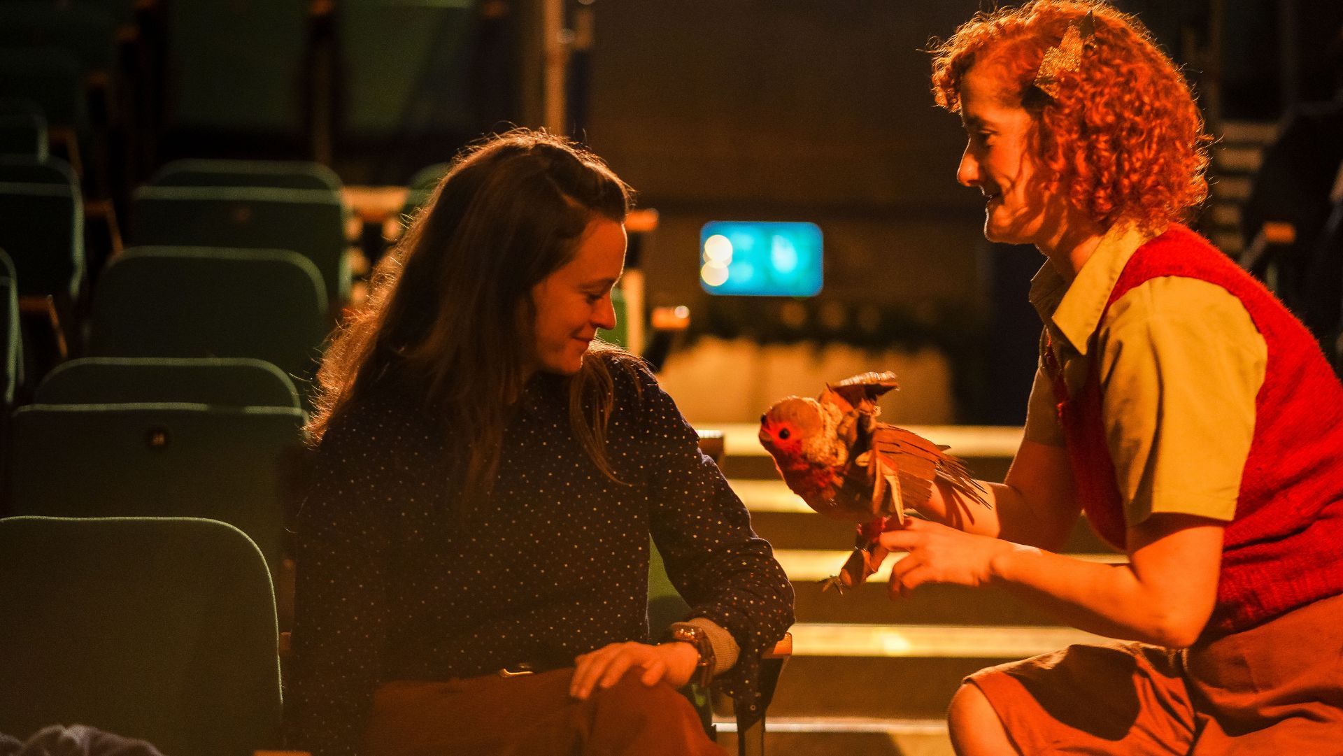 An audience member in a dark, polka-dotted blouse smiles while interacting with a performer holding a puppet of a robin. The performer, wearing a red knitted vest and beige shirt, leans forward engagingly. The setting is a theatre with rows of green seats in the background, illuminated by warm stage lighting.