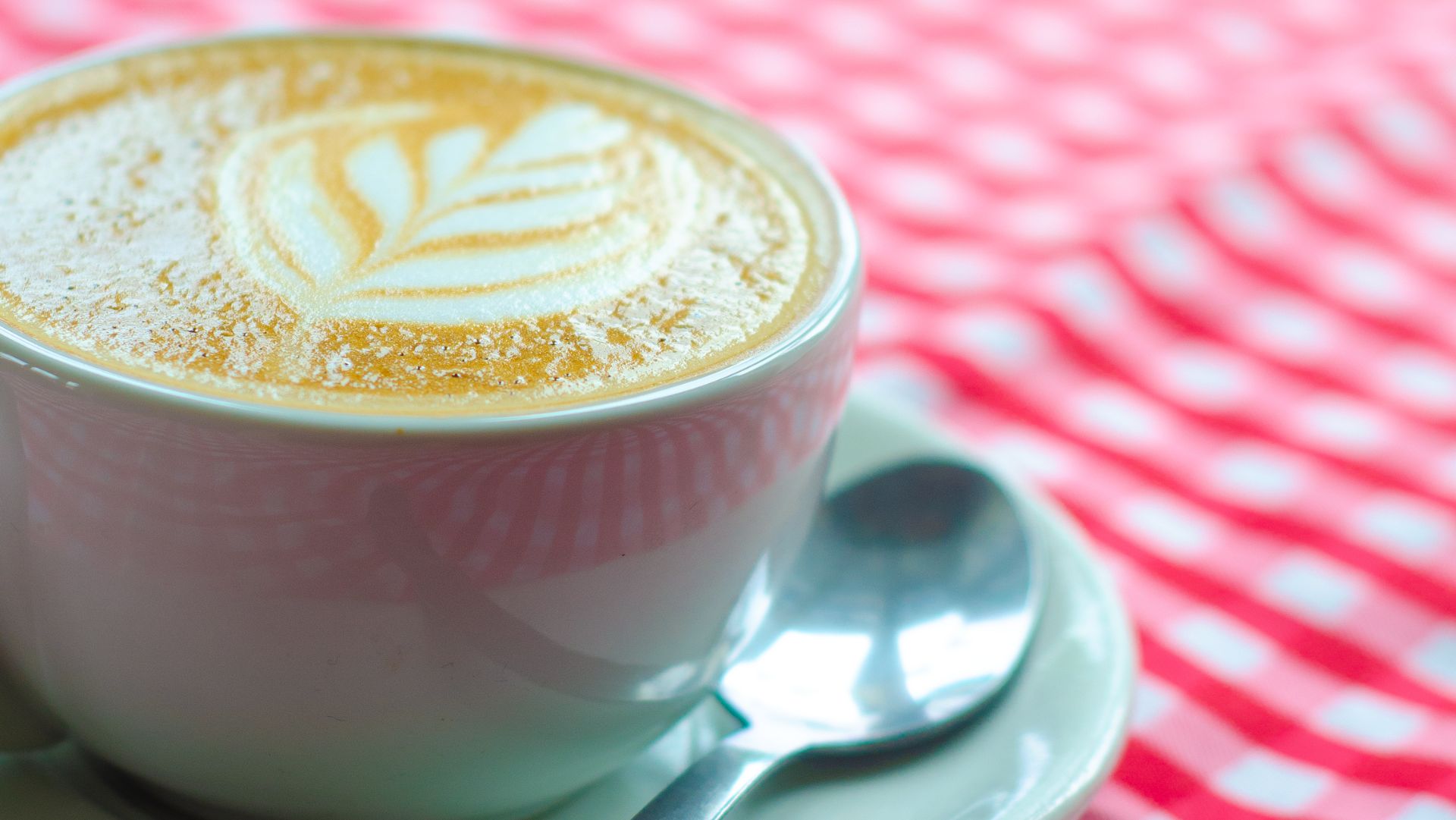 A white mug filled with coffee with pretty coffee art on top sat on a saucer with a spoon on the side. Sat on top of a red and white gingham tablecloth.