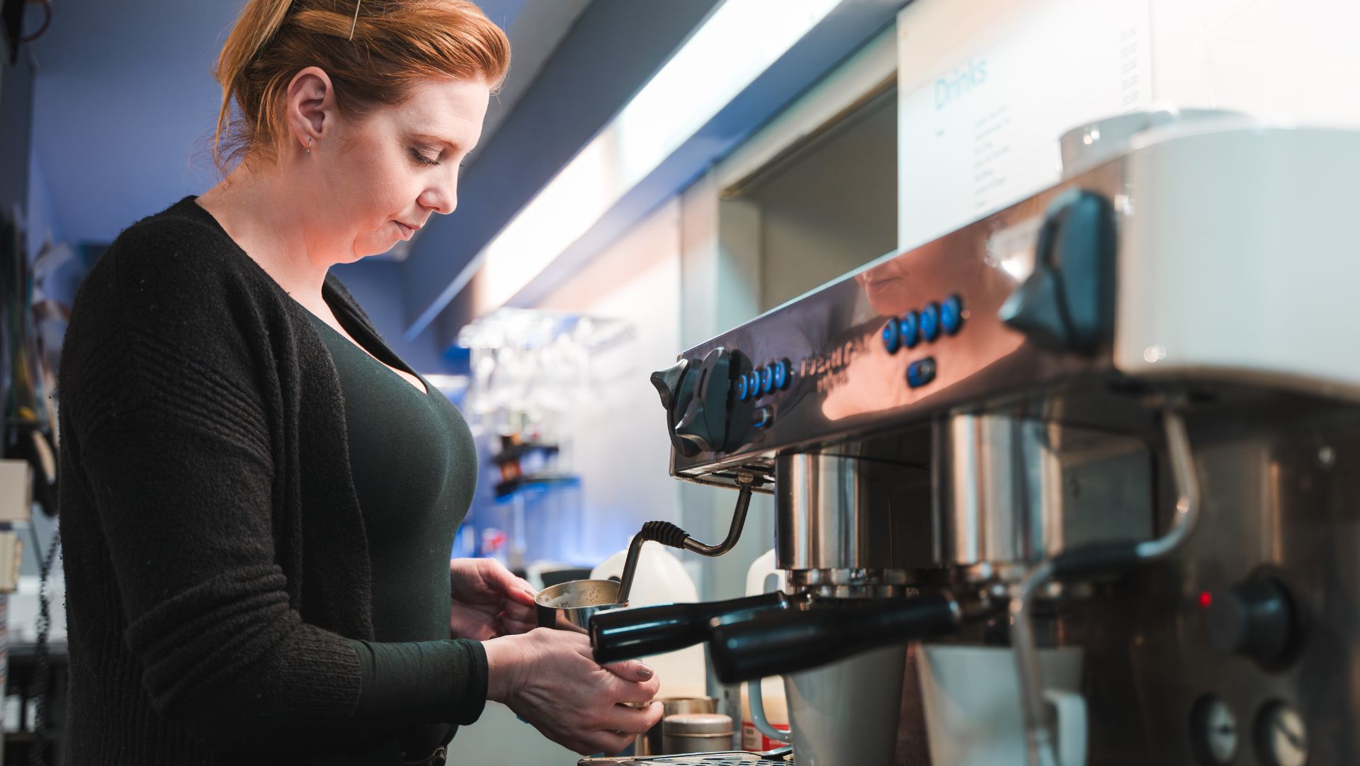 A woman with blonde hair wearing a black cardigan and top is using a coffee machine to make a coffee for a customer