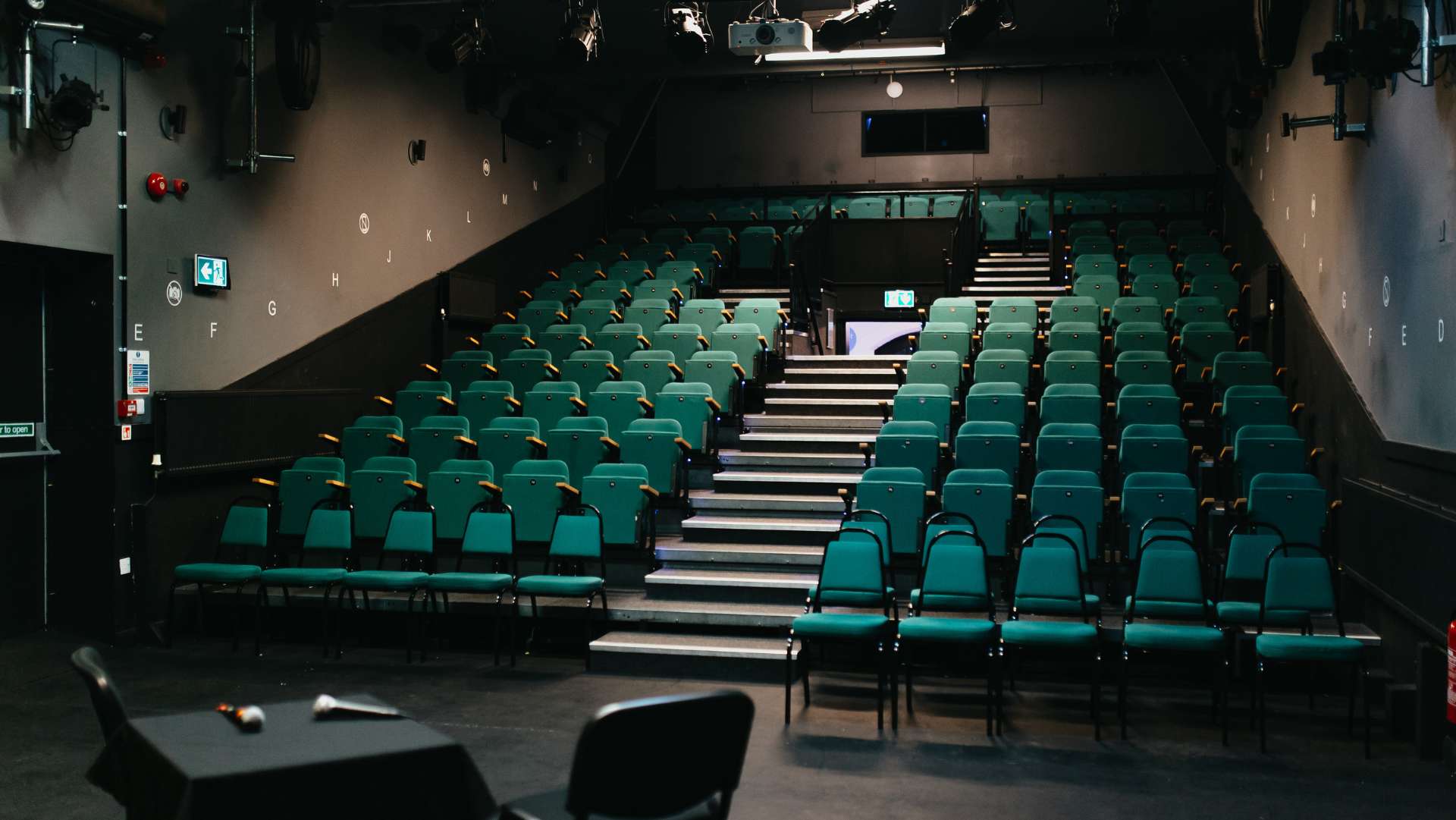 An empty theatre with a small capacity of 134 seats with teal chairs and stairs going down the middle towards the stage.