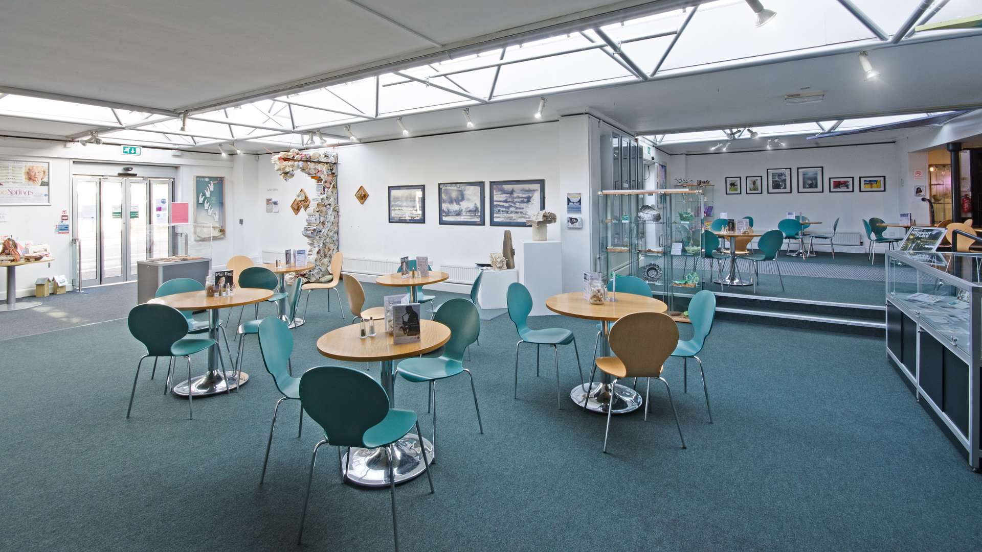 A bright and open play foyer space with tables and chairs positioned around the room. The floor is a teal carpet and the walls are white. It is a comfortable space with artworks hung up on the walls.