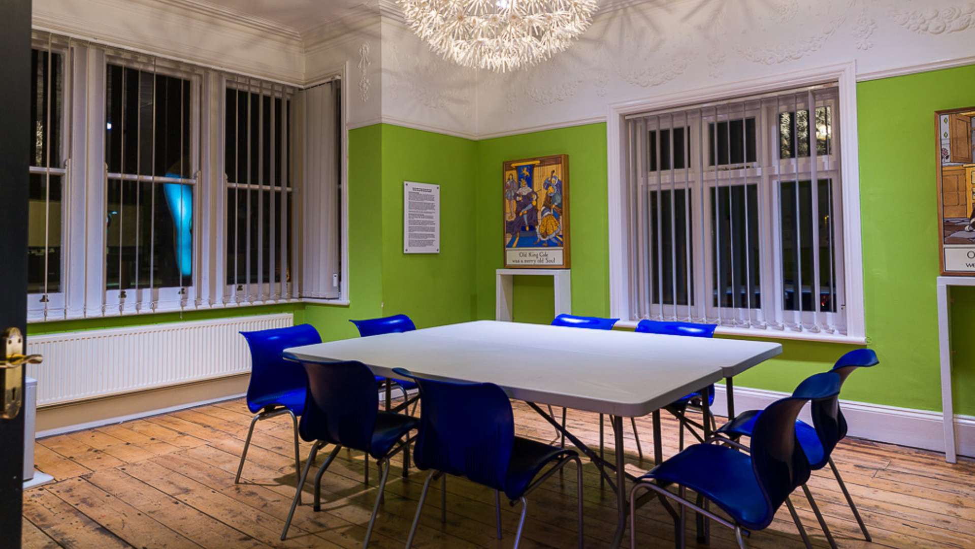 A brightly lit room with lime green walls, wooden flooring, and a rectangular table surrounded by blue chairs. The room features large windows with vertical blinds, and a decorative light fixture hangs from the ceiling. Framed artwork and informational plaques are mounted on the walls.