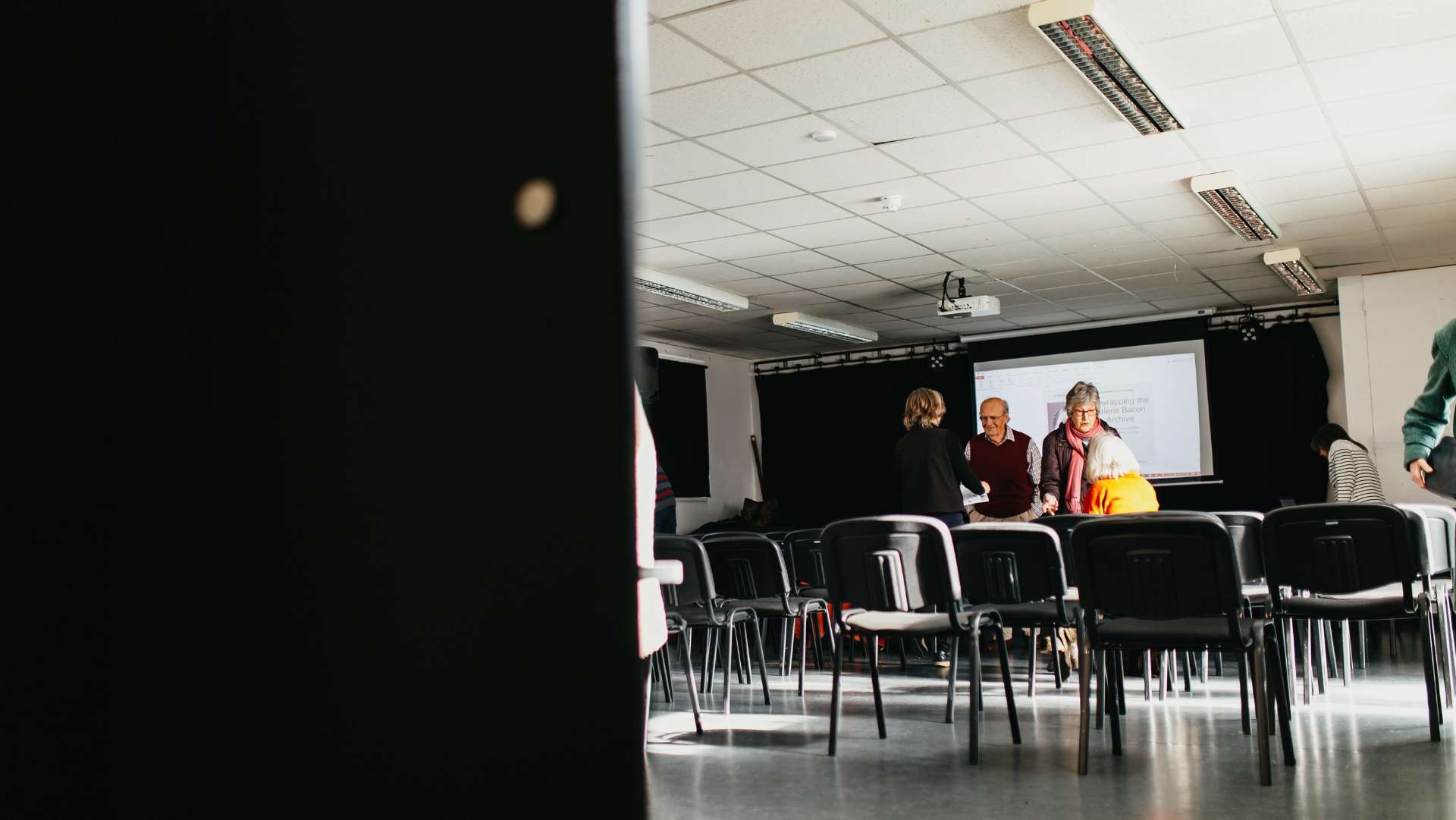 A small group of people gather in a bright, modern room with a projector screen at the front. The chairs are arranged in rows, and the atmosphere suggests a casual meeting or workshop. Fluorescent lights line the ceiling, and sunlight streams through the windows.