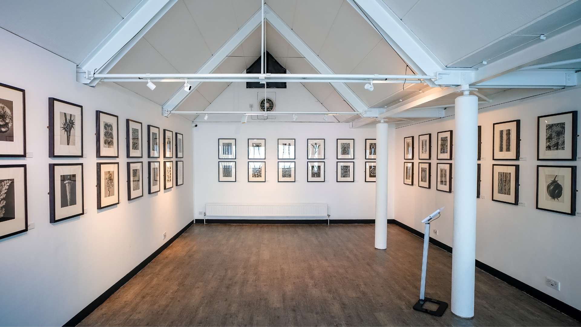 A well-lit gallery space with high, vaulted ceilings and white walls displaying an array of framed black-and-white artwork. The floor has a wooden finish, and track lighting highlights the art pieces. A white column and a small adjustable stand are visible in the room, adding to the minimalist design. The overall atmosphere is clean and modern, ideal for exhibitions.