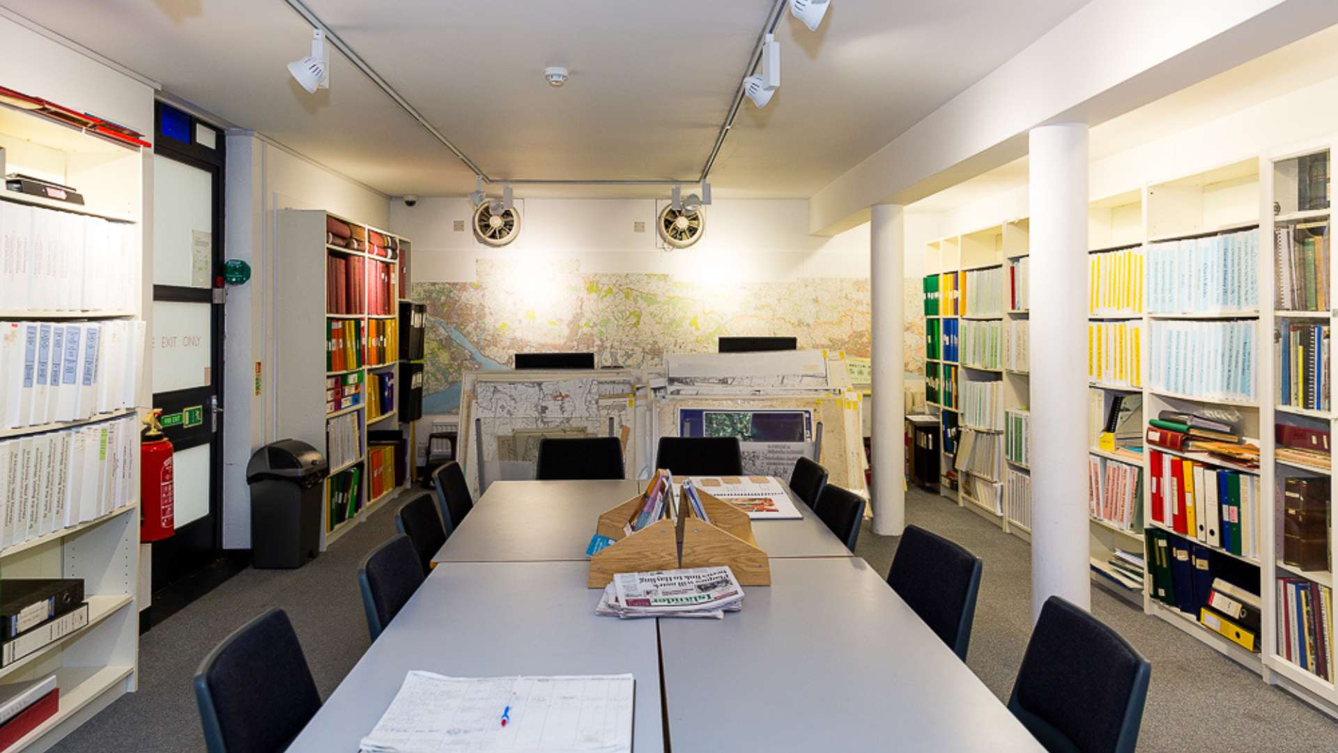 A warm white room with a white table in the middle of the room. There are shelves alongside the side walls filled with folders of archives and records of Havant's history. There are also two desktop computers at the back of the room.