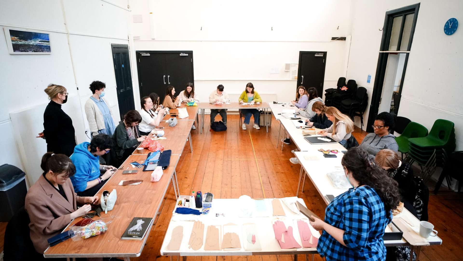 A large room with a wooden floor and white walls. Going around the room are brown fold-out tables where there are people sitting in chairs taking part in a workshop making gloves.