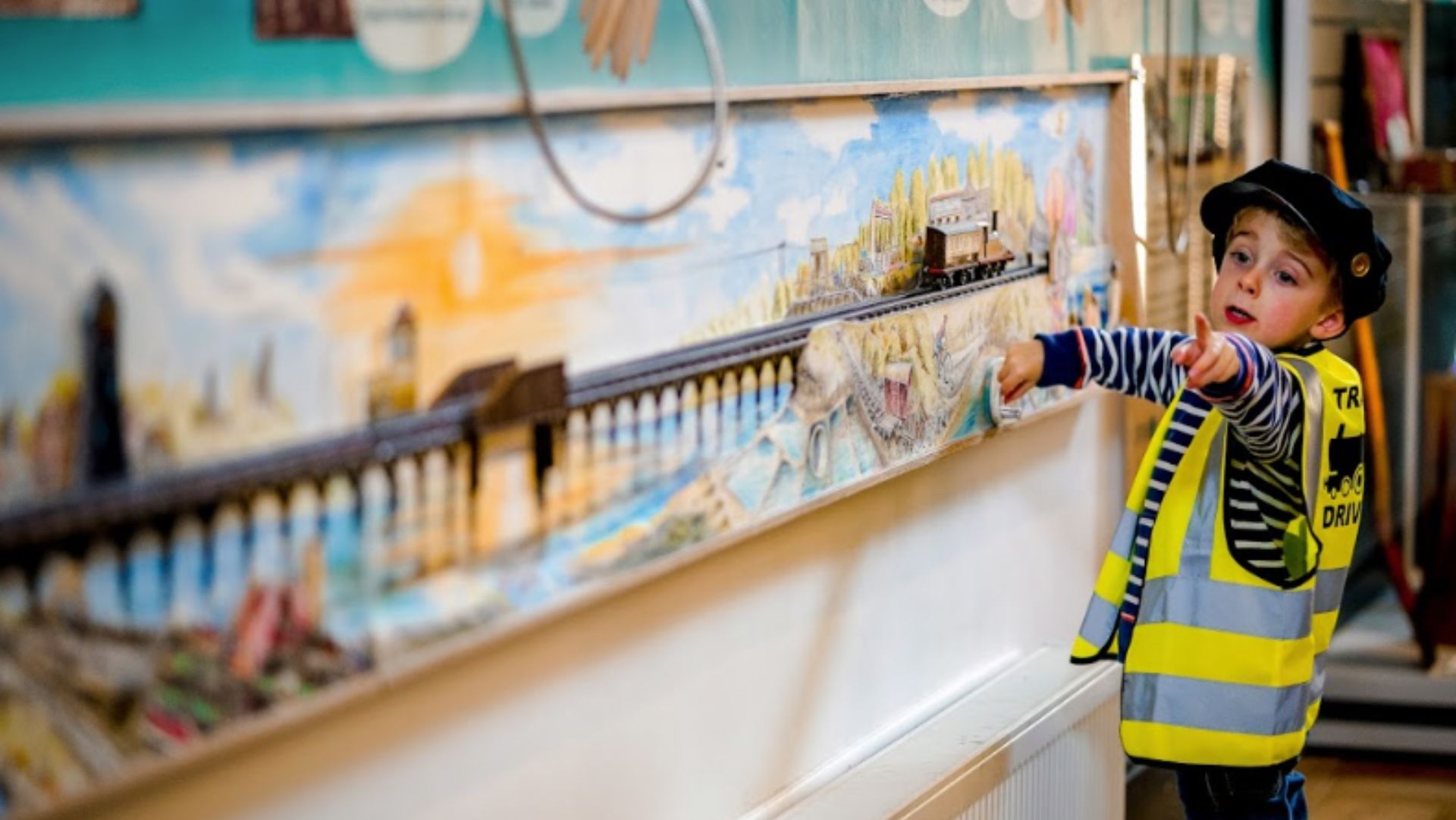 A young boy wearing a stripe t-shirt with a high vis over the top is pointing whilst holding a turn handle in front of a railway model