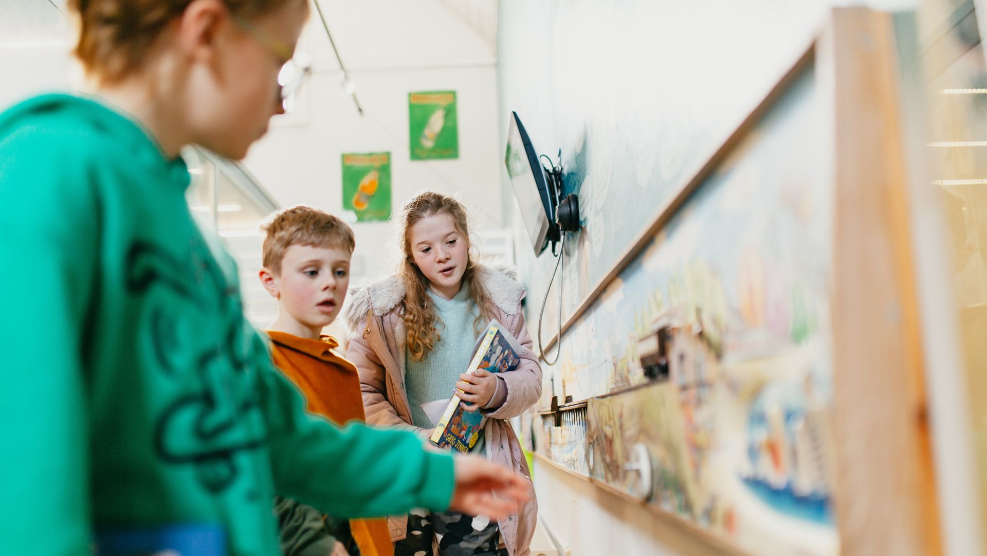 Three children engaging with a museum exhibit, looking closely at a colourful, interactive display mounted on the wall. One child in a pink coat holds a book while observing intently, accompanied by another child in an orange jumper and a third child partially visible in a green jumper. The setting is bright and inviting, with additional posters visible in the background.