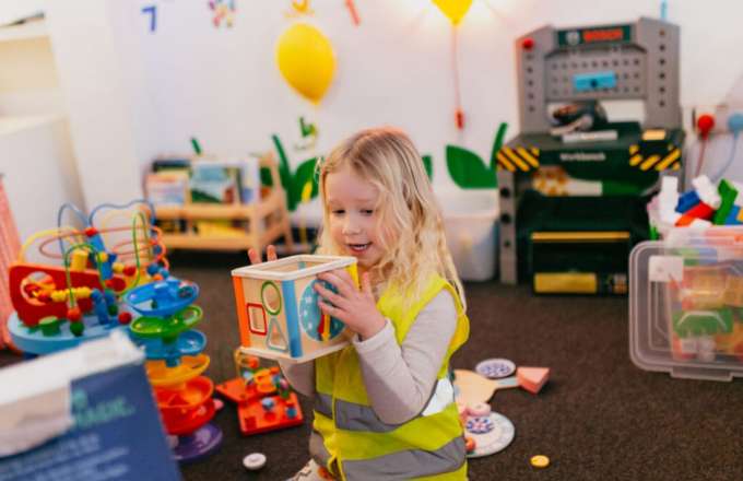 A young girl with blonde hair wearing a white top with a high vis jacket on is holding a puzzle block for children. She is stood in our family play space surrounded by various toys a child could play with