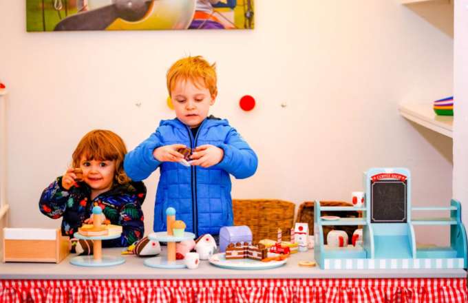 Two children, one a younger girl with brown hair wearing a black coat and an older boy with ginger hair in a blue coat stand behind a counter with a red and white gingham cover. With various toy pieces of food and a cafe counter to look like they are running a make-believe café.