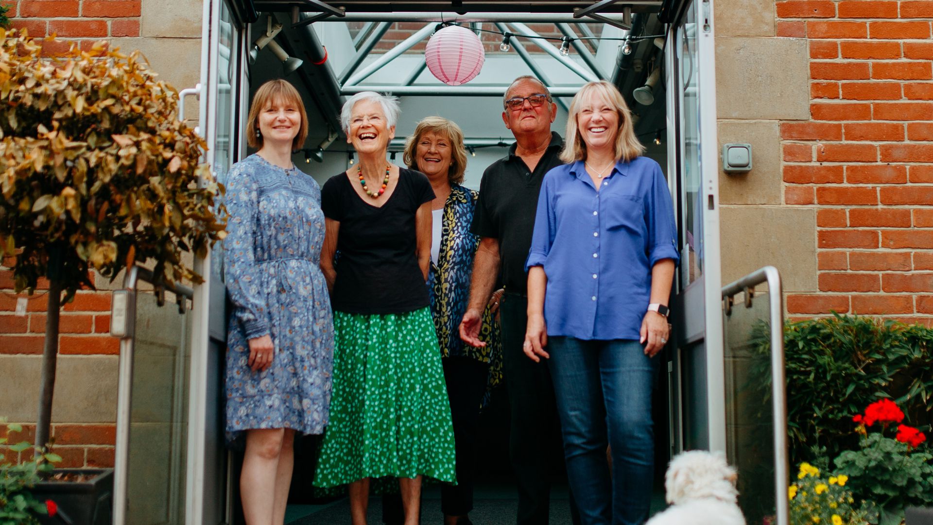 Five cheerful adults stand together at the entrance of a building with a glass canopy overhead. They are smiling and appear to be enjoying the moment. The group includes three women wearing colourful clothing, an older man in a dark polo shirt and sunglasses, and another woman in a vibrant blue blouse. Brick walls and a potted shrub frame the doorway.